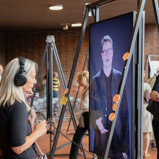 Eine Besucherin lauscht dem Interview mit Jens Hendrix, Baudezernent in Hattingen zu Kirchentransformationen. Ausstellungseröffnung „Kirchen als Vierte Orte – Perspektiven des Wandels“, 1.9.2024, Heilig-Geist-Kirche, Essen.