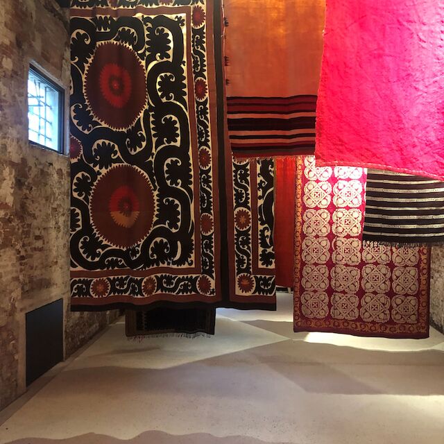 Die Installation auf der linken Seite in der Präsentation von „Keep Cool“: riesige, strahlend bunte, an Querstreben aufgehängte Teppiche, Decken und Tücher bewegen sich langsam vor und zurück durch den Raum.